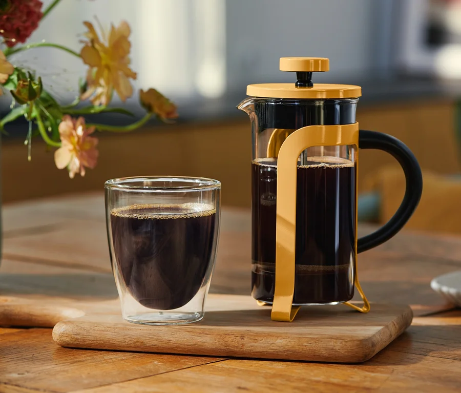 Çift cidarlı bir bardak siyah kahve ve kahve dolu sarı bir French Press, ahşap bir tahta üzerinde duruyor. Arka planda çiçekli bir vazo ve kurabiyeli küçük bir tabak var.