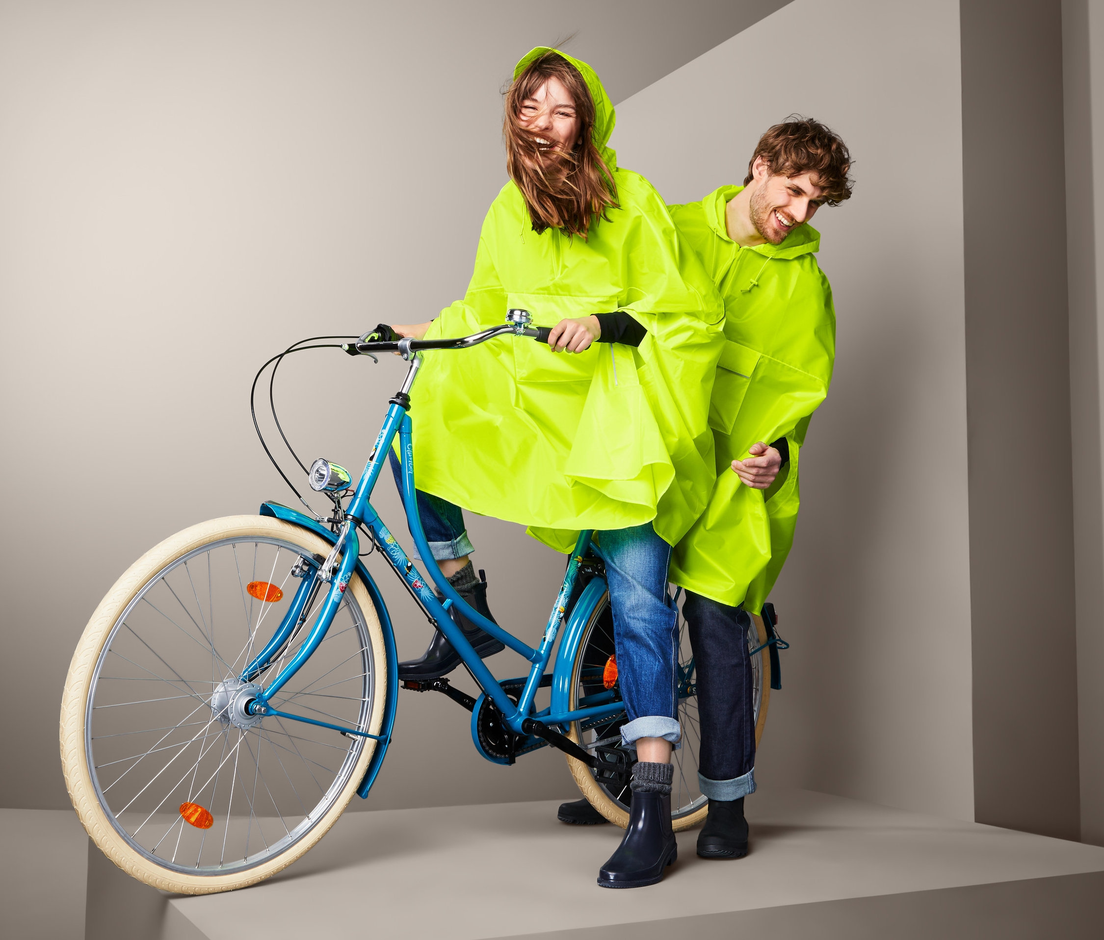 Kadın mavi bir bisiklete biniyor, arkasında bir adam var, her ikisi de sarı PANÇO YAĞMURLUK giyiyor.