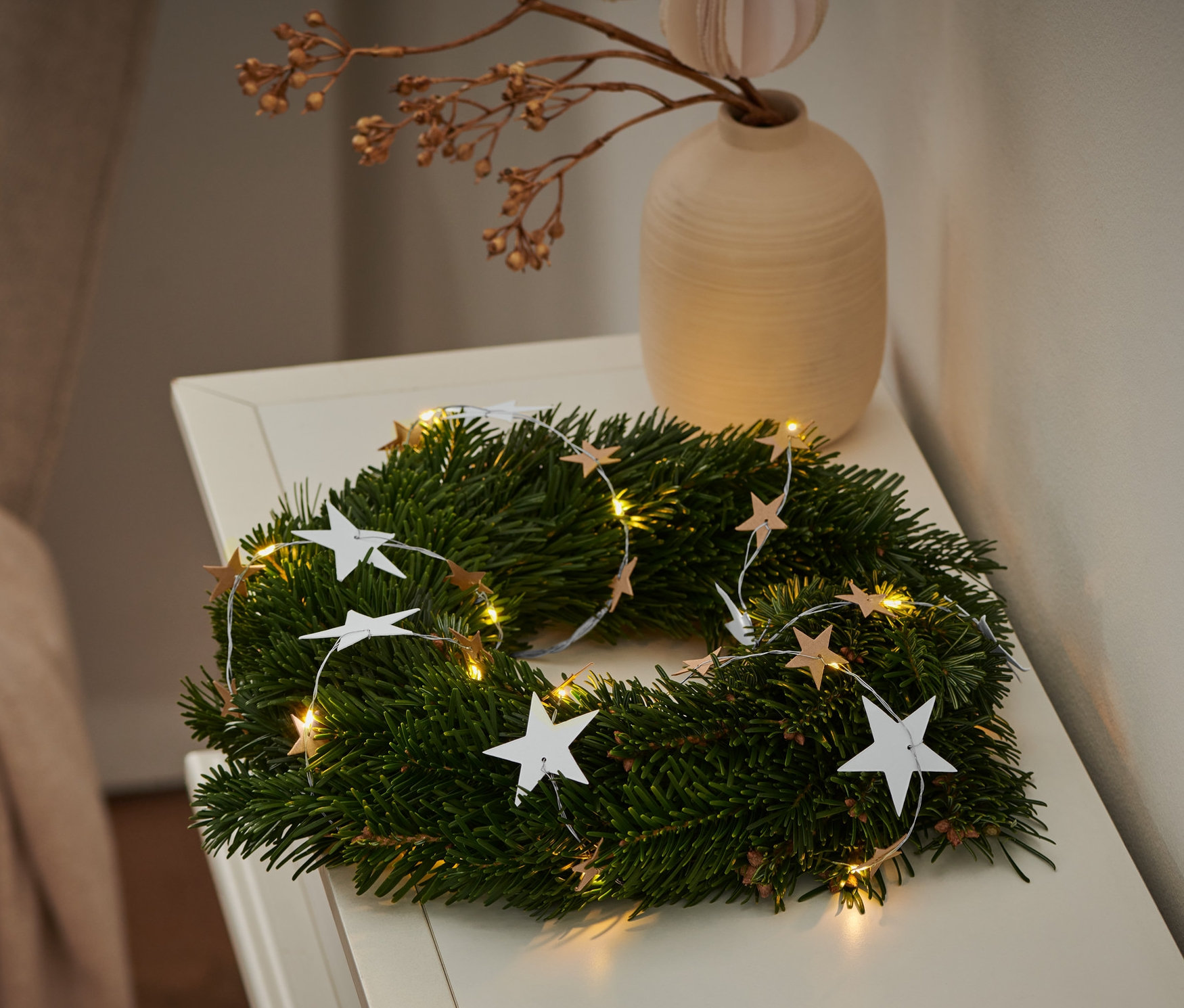Beyaz bir masa üzerinde LED Işık Zinciri »Kağıt Yıldızlar« olan yeşil bir çelenk. Çelenkin yanında bir tabak kurabiye var.