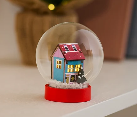 Mavi evi ve kırmızı tabanı olan LED Kar Küresi bir rafta duruyor.