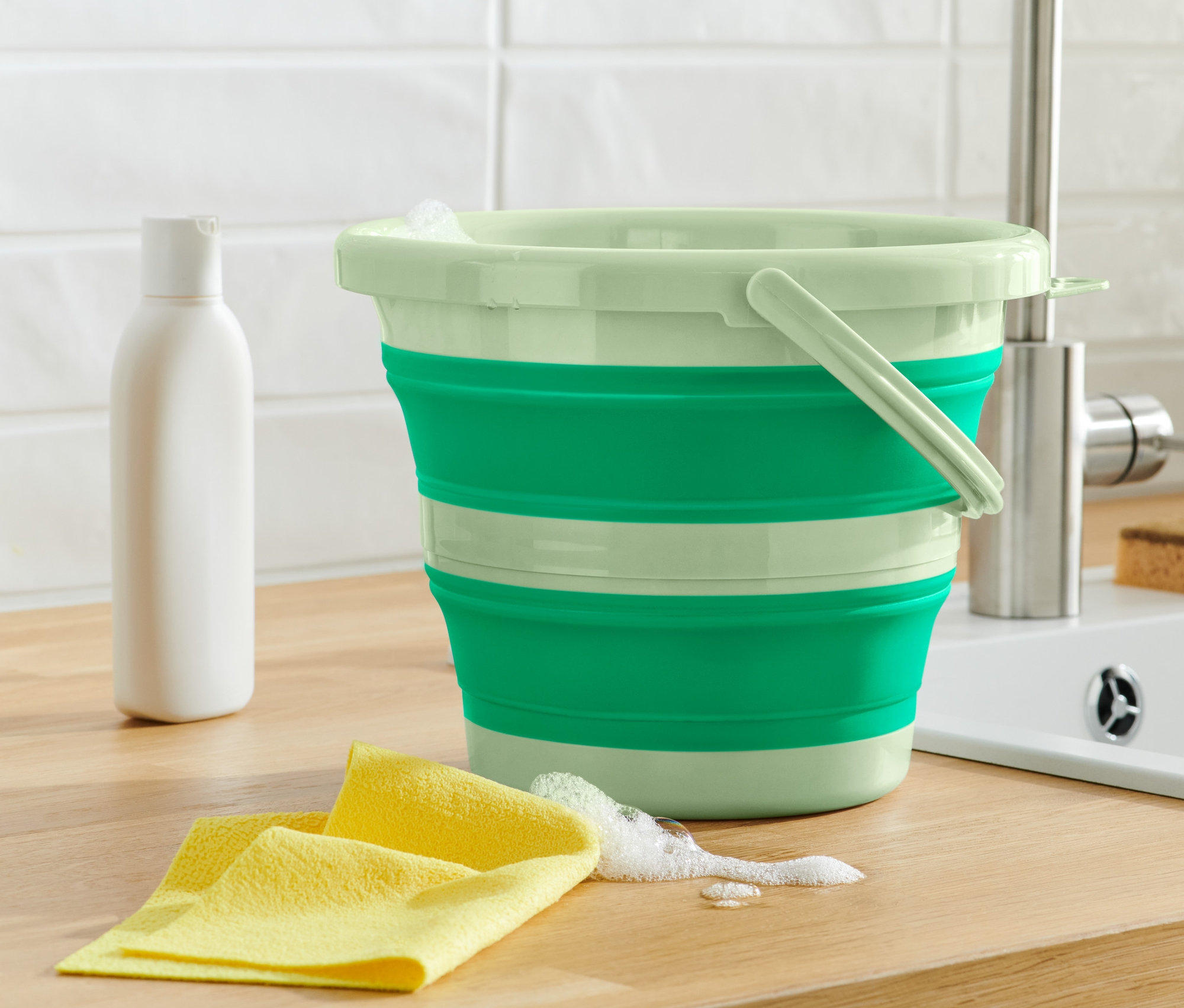 Yeşil katlanabilir temizlik kovası, sarı bez ve beyaz şişenin yanında ahşap tezgah üzerinde duruyor.