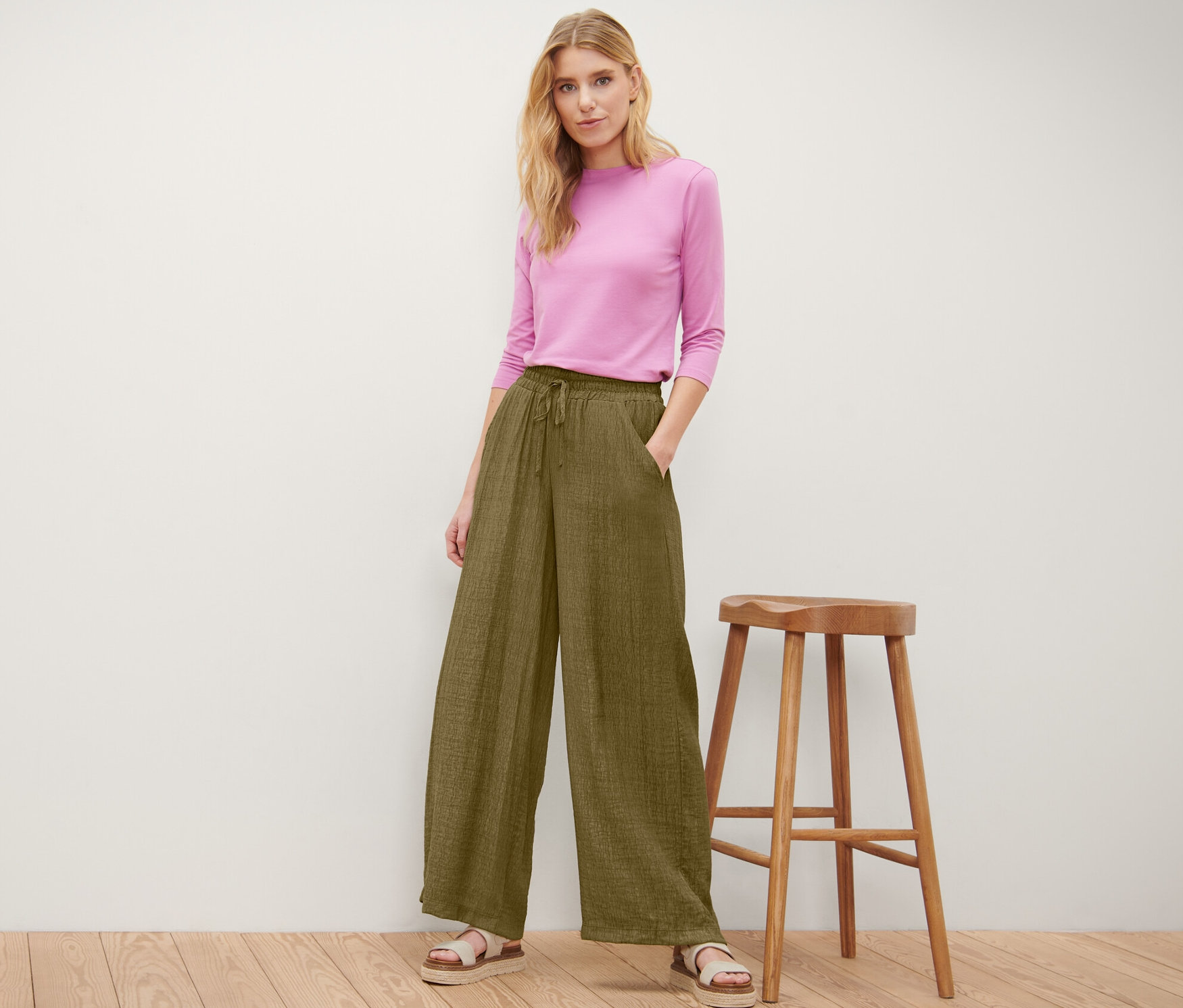 Kadın, zeytin yeşili eşofman pantolon ve pembe üstle ahşap taburenin yanında poz veriyor.