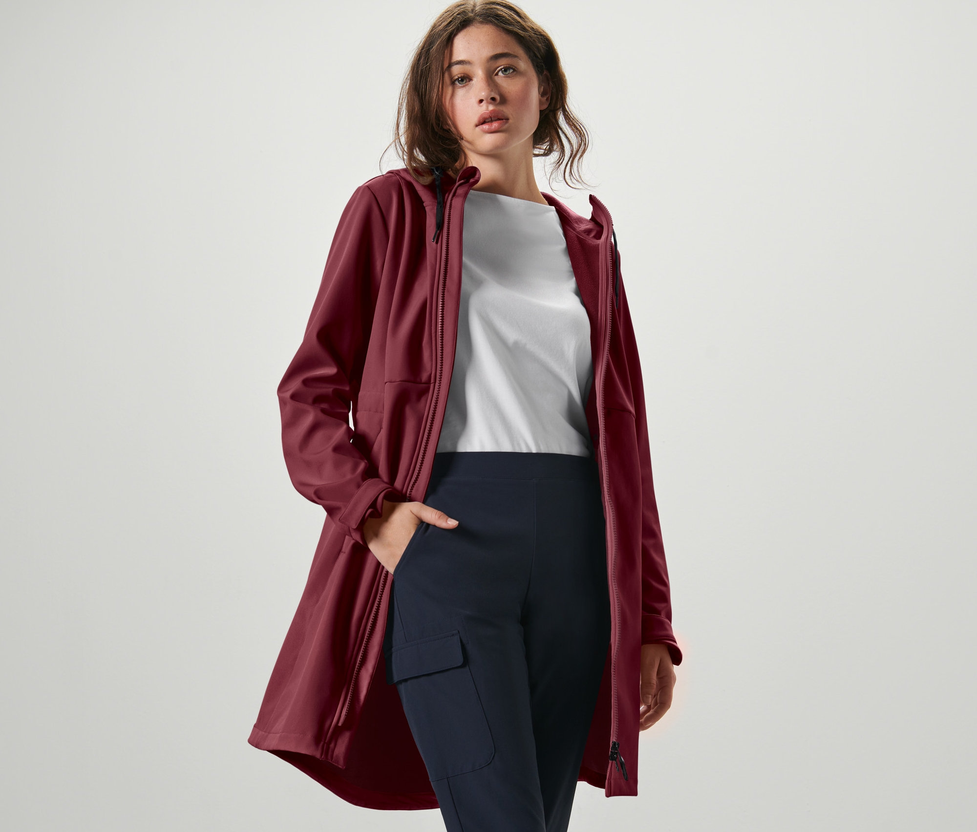 Kadın şarap kırmızısı softshell palto, beyaz tişört ve lacivert pantolonla poz veriyor.