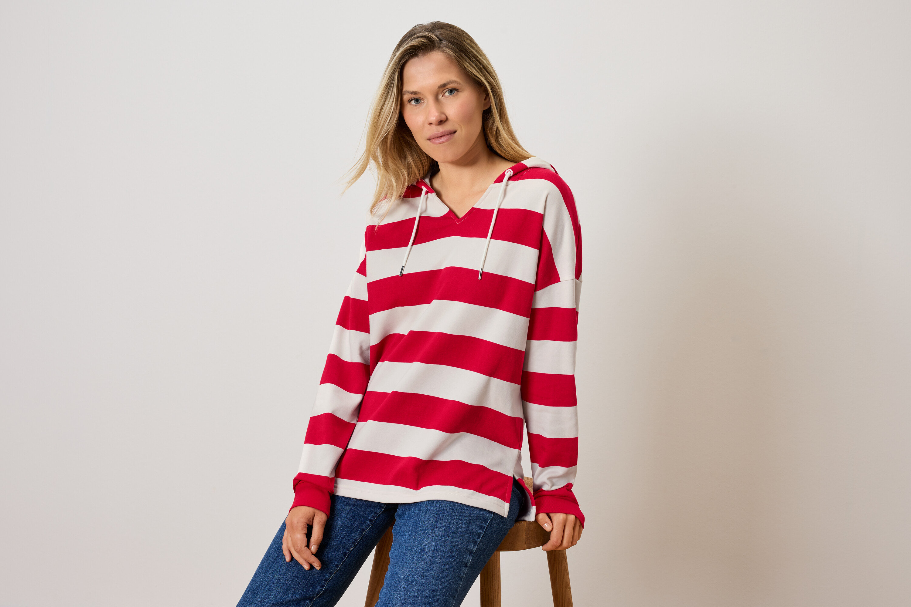 Bir kadın taburede oturuyor, çizgili kapüşonlu bir sweatshirt ve kot pantolon giyiyor.