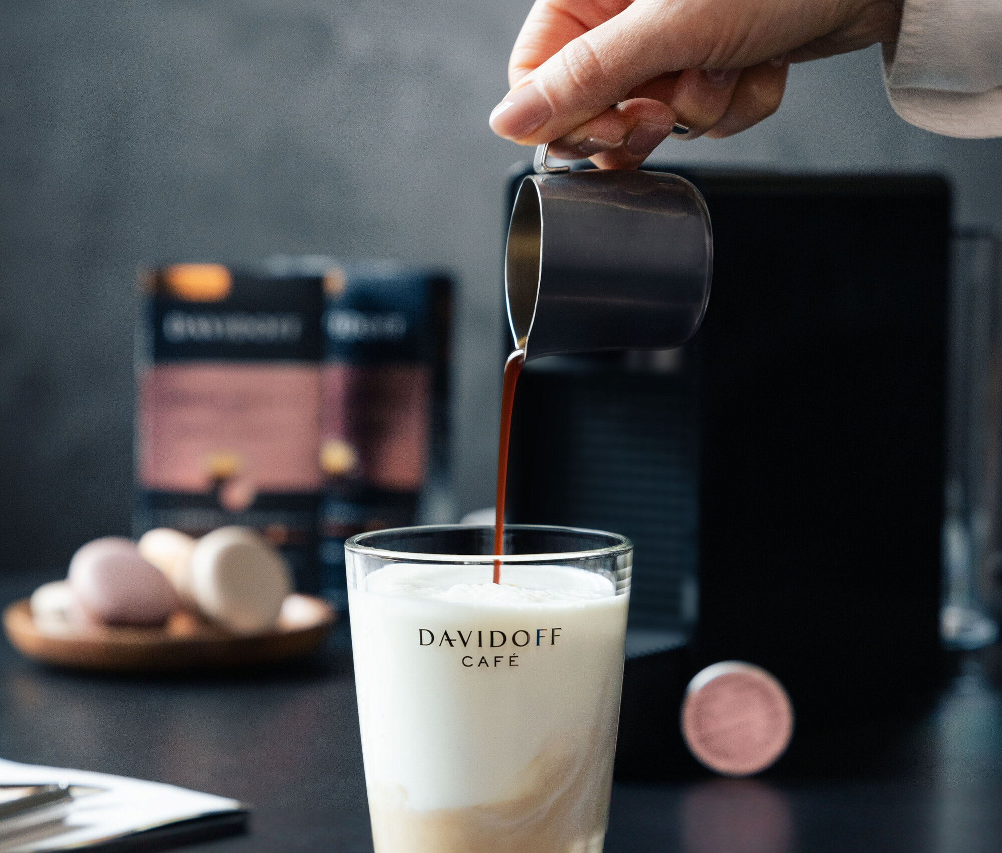 Bir el sütlü bir bardağa kahve döküyor. Arka planda bir kahve makinesi ve bir paket DAVIDOFF CAFÉ Crema Intense bulunuyor.
