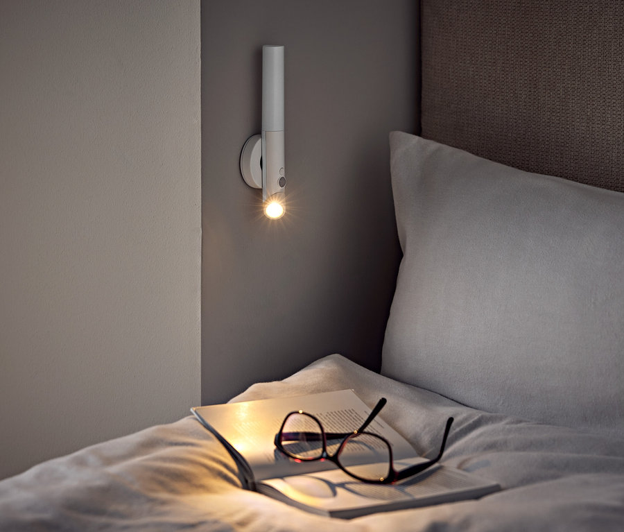 Yatakta, bir duvar lambasının altında bir gözlük ve açık bir kitap duruyor.