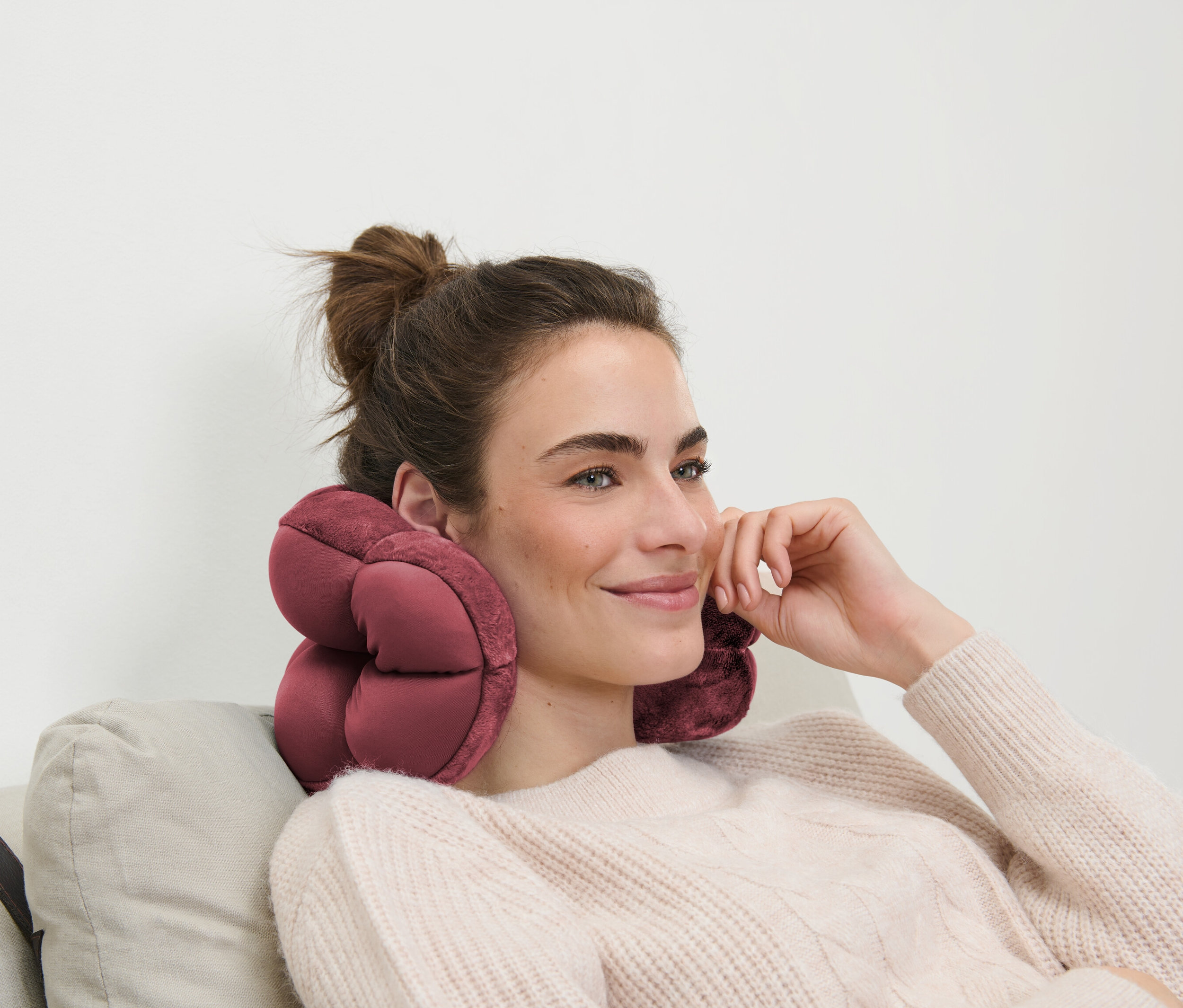 Kahverengi saçlı bir kadın, boynunda pembe bir seyahat yastığıyla bir kanepede oturuyor.