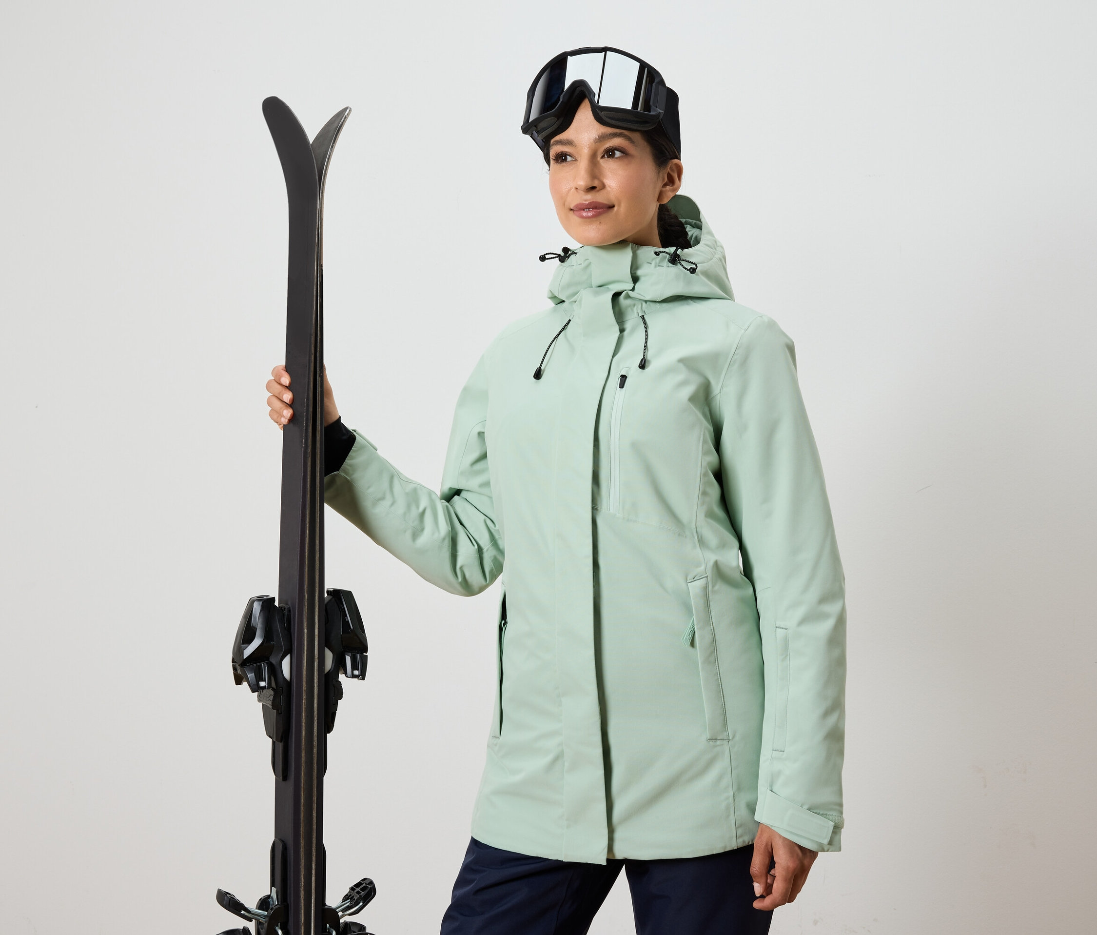 Kadın kayak tutuyor, kayak gözlüğü ve ceket giyiyor.