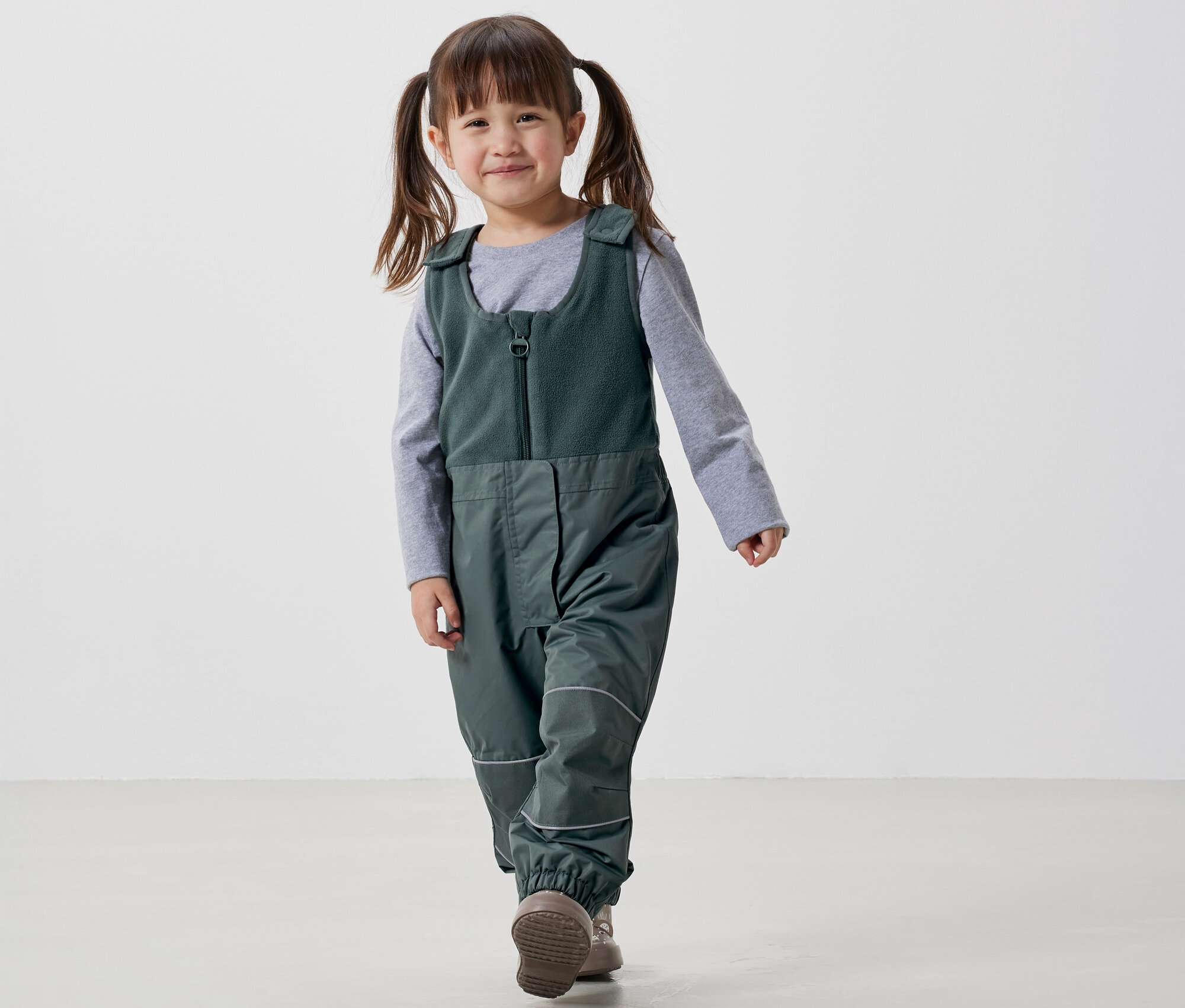 Gülümseyen at kuyruklu küçük kız, beyaz bir arka planda yeşil salopet ve gri tişörtle yürüyor.