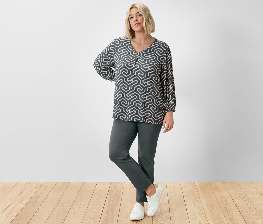 Bir kadın stüdyoda koyu yeşil streç pantolon ve kırışık görünümlü bluzla poz veriyor.