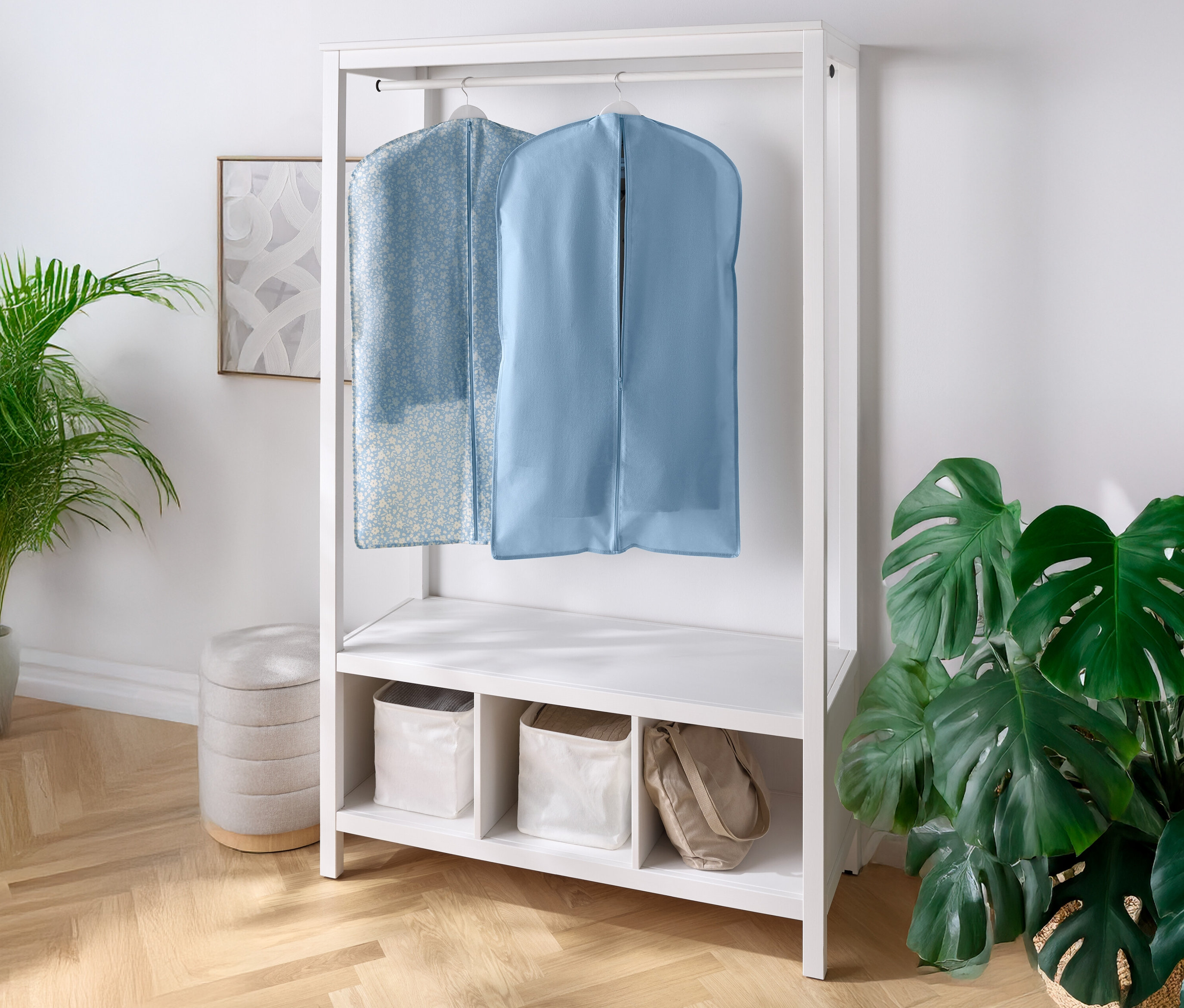 İki elbise kılıfı, altında rafları ve bölmeleri olan beyaz bir vestiyerde asılı duruyor. Sol ve sağ tarafta birer ev bitkisi bulunuyor.