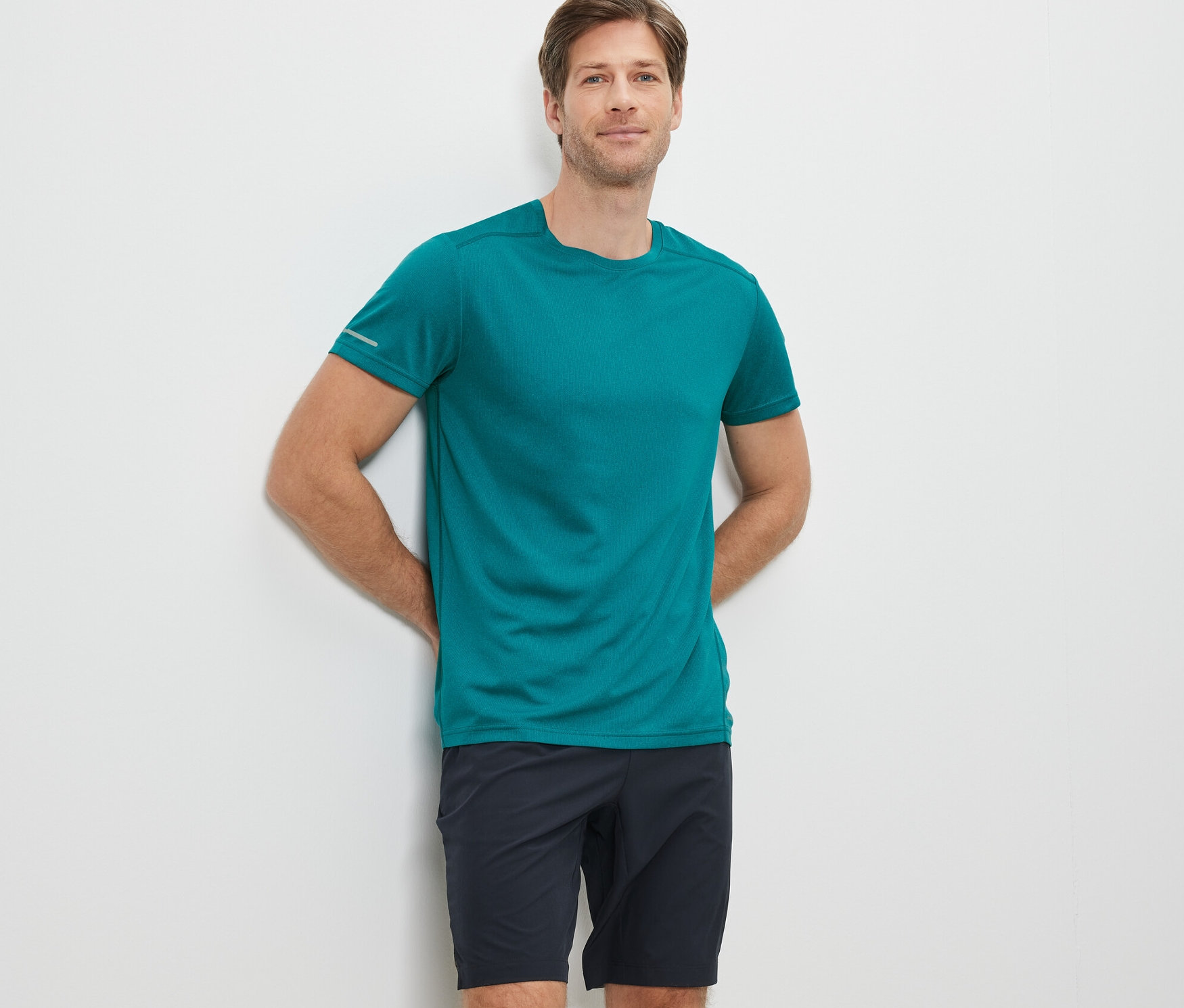 Beyaz duvara yaslanmış bir adam, turkuaz fonksiyonel tişört ve siyah şort giyiyor.