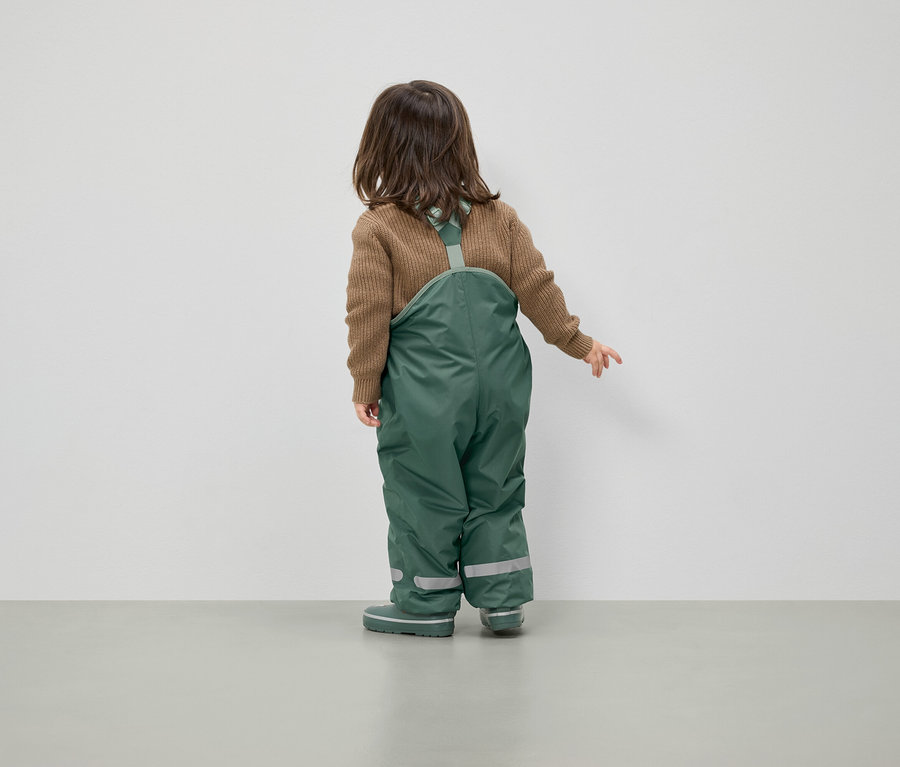 Kahverengi kazak ve yeşil salopet giyen bir çocuk kameraya arkası dönük duruyor.