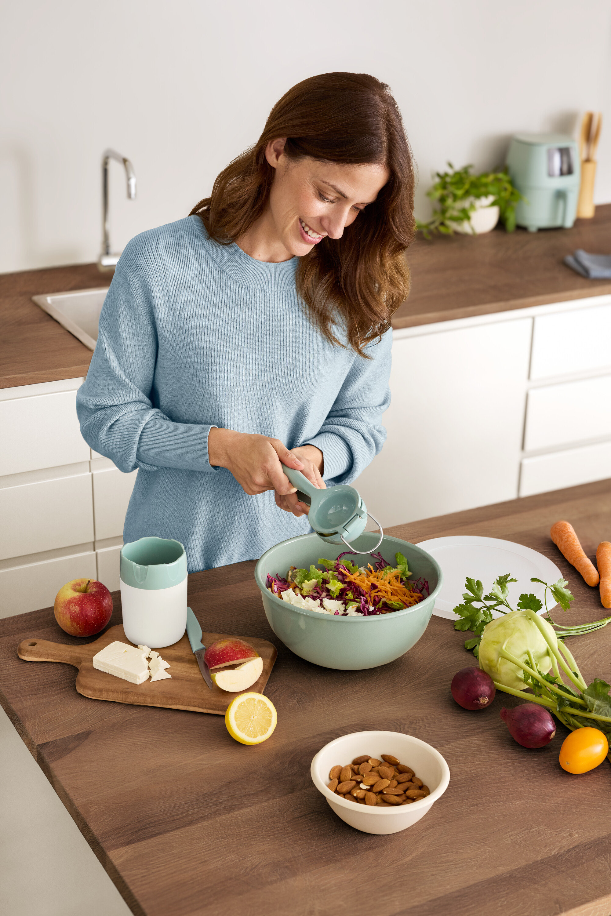 Kadın salata kasesine limon sıkıyor. Masada elma, peynir, badem ve sebzeler var.