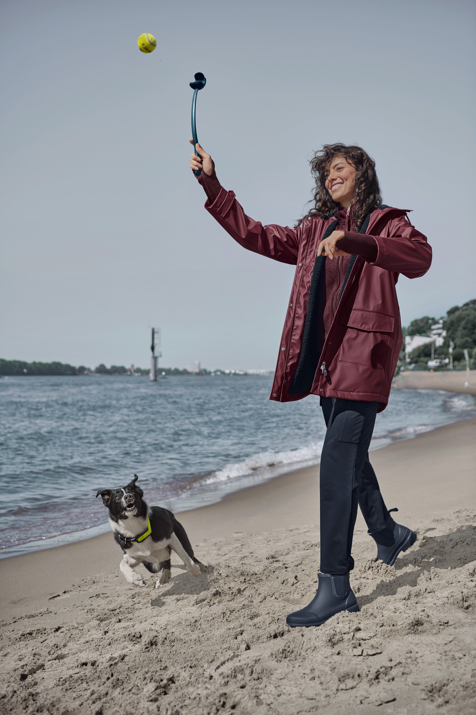 Sahilde bir kadın, top fırlatıcı ile köpeğe top atıyor. Üzerinde şarap kırmızısı örgü polar mont, teddy yağmurluk ve lacivert kargo cepli jogger pantolon var.