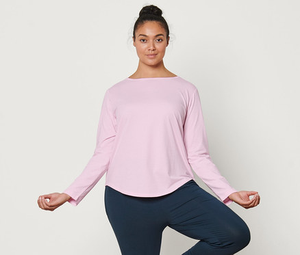 Kadın yoga matı üzerinde ağaç duruşunda duruyor. Üzerinde açık pembe organik pamuklu uzun kollu spor tişörtü ve yoga pantolonu var.