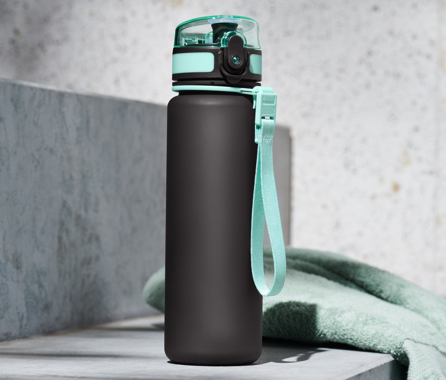 Turkuaz kapaklı ve askılı siyah matara, turkuaz bir havlunun yanında beton bir duvarın üzerinde duruyor.