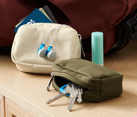 İki adet mini seyahat düzenleme çantası, kulaklık ve dudak balsamı bir sırt çantası ve vazonun yanında bir komodinin üzerinde duruyor.