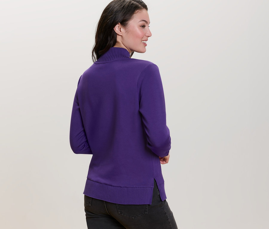 Kadın omzunun üzerinden bakıyor, üzerinde mor, yarım balıkçı yaka sweatshirt var.