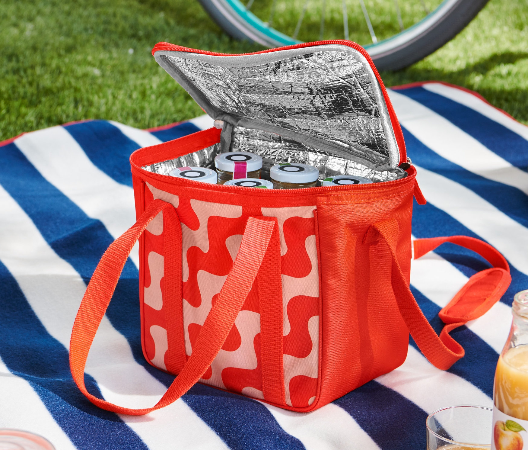 Açık, kırmızı ve beyaz desenli küçük bir soğutucu çanta, mavi ve beyaz çizgili bir örtü üzerinde duruyor ve içi kavanozlarla dolu. Arka planda bir bisiklet ve kask görünüyor.