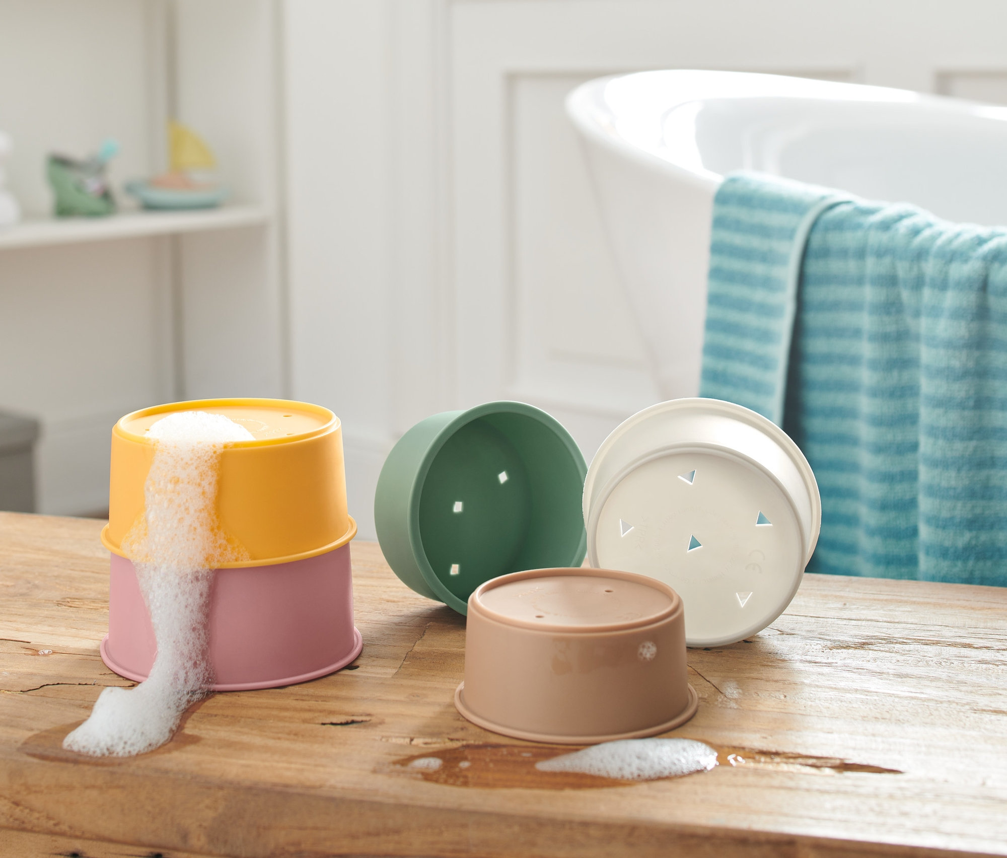 Ahşap bir masa üzerinde iç içe geçmeli banyo oyun kapları bulunuyor, ikisi üst üste yerleştirilmiş ve üstteki kaptan köpük taşıyor.