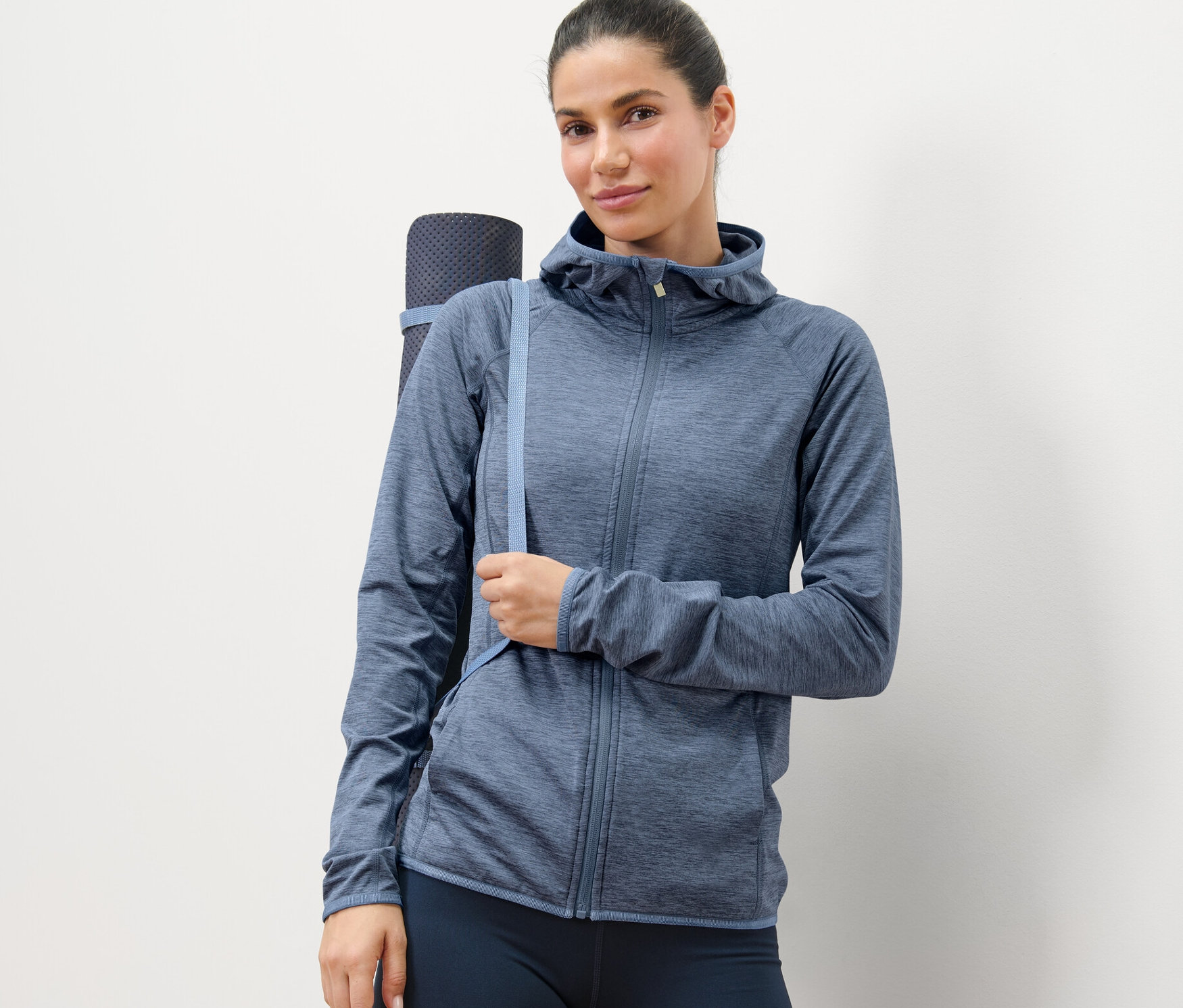 Kadın kırçıllı mavi spor mont giyiyor ve yoga matı taşıyor.