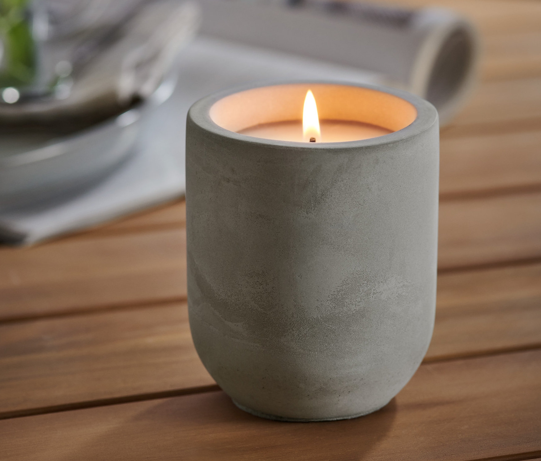 Ahşap bir masa üzerinde beton bir kap içinde yanan bir Limon Kokulu Mum bulunmaktadır. Arka planda bitki ve tabak bulunan bir bardak.