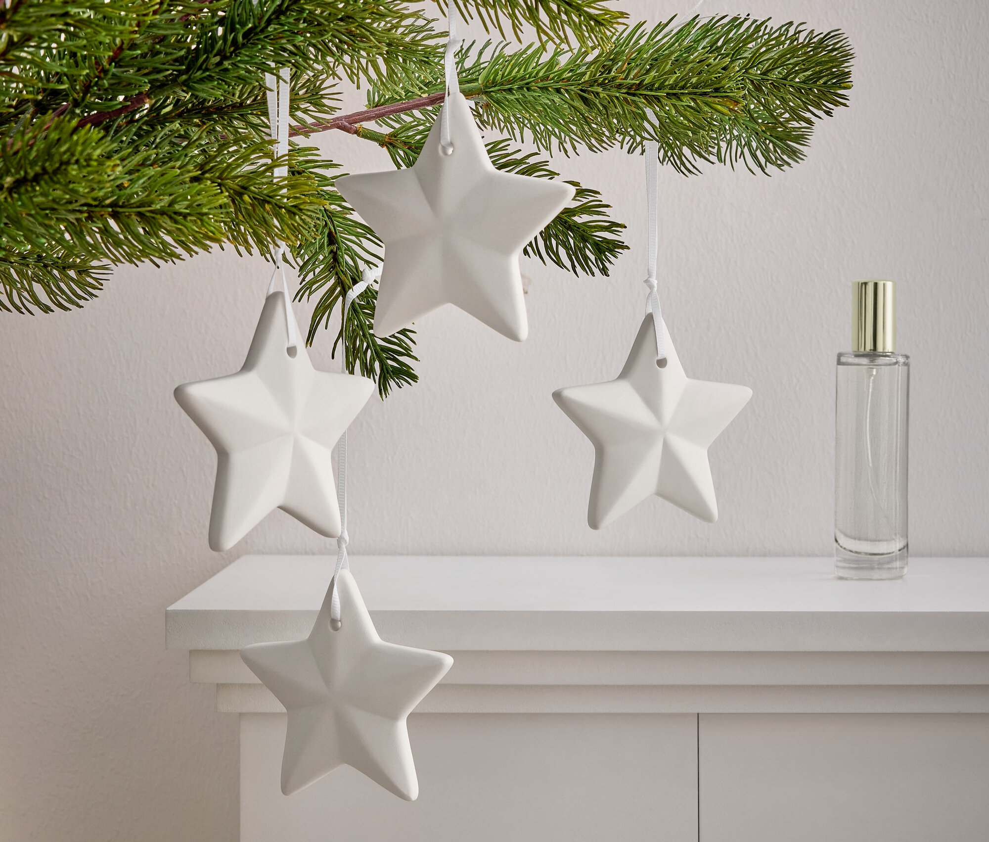 Beyaz bir dolabın üzerinde dört adet beyaz yıldızın asılı olduğu iğne yapraklı bir dal. Yıldızların yanında bir parfüm şişesi duruyor.