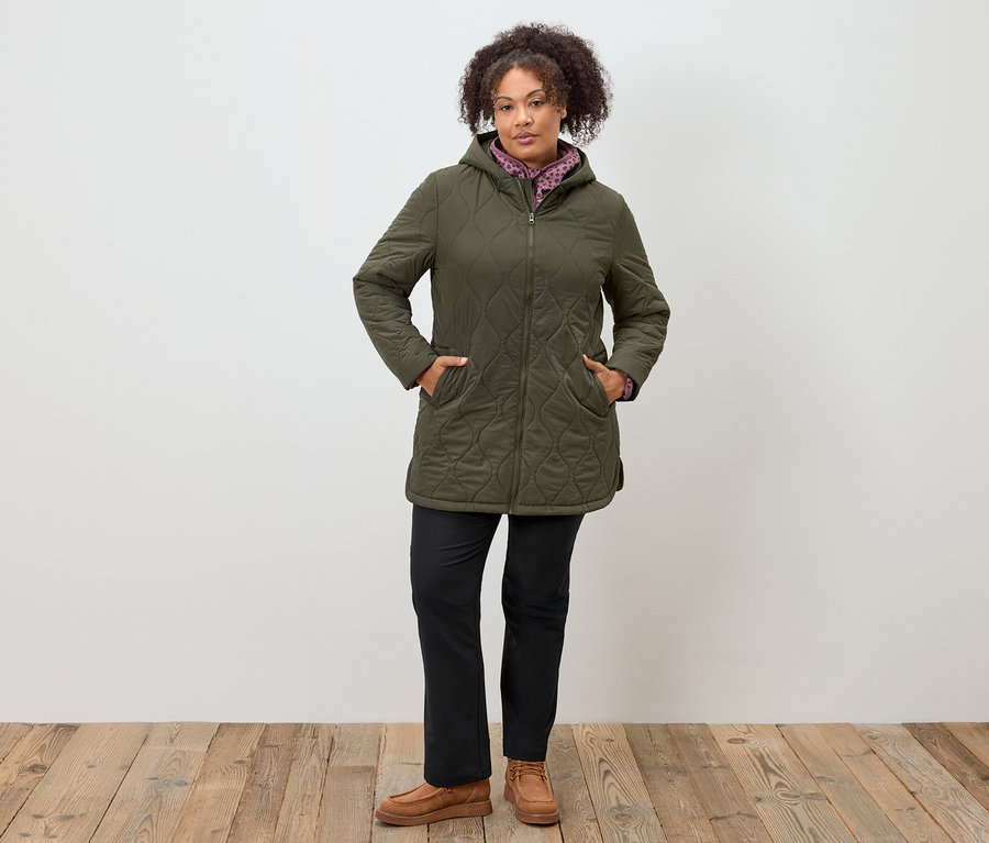 Kadın ayakta duruyor, elleri zeytin yeşili kapüşonlu kapitone ceketinin cebinde, siyah pantolon ve kahverengi mokasen giyiyor.