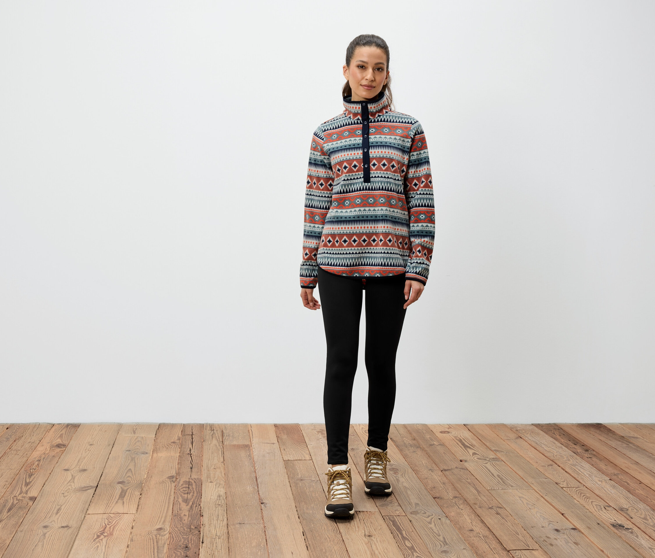 Bir kadın ahşap zeminde duruyor, desenli bir sweatshirt, siyah tayt ve kahverengi botlar giyiyor.