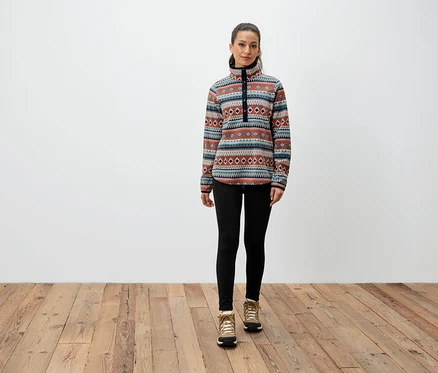 Bir kadın ahşap zeminde duruyor, desenli bir sweatshirt, siyah tayt ve kahverengi botlar giyiyor.