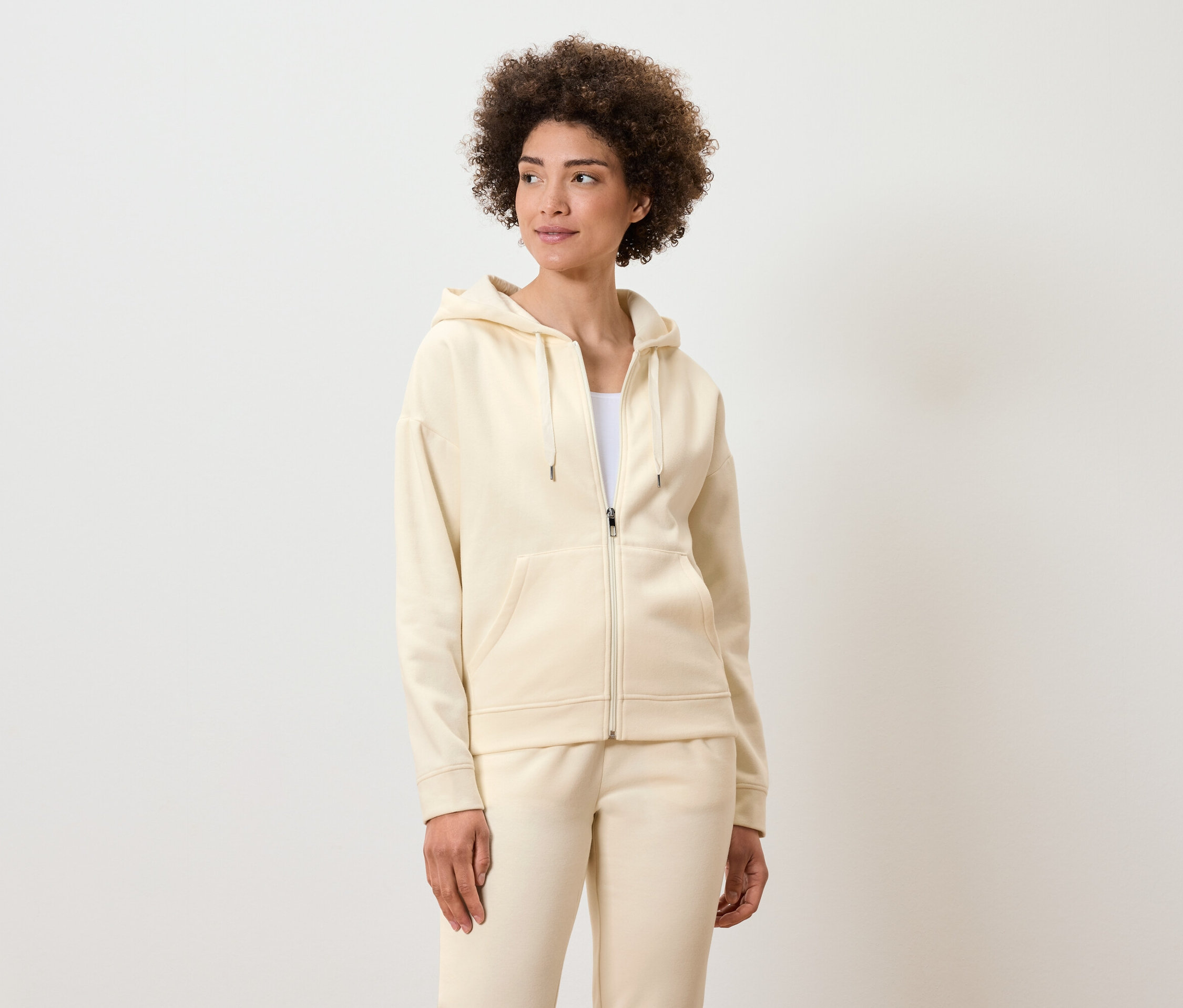 Kıvırcık saçlı kadın, krem rengi, fermuarlı kapüşonlu sweatshirt ve eşofman altıyla poz veriyor.