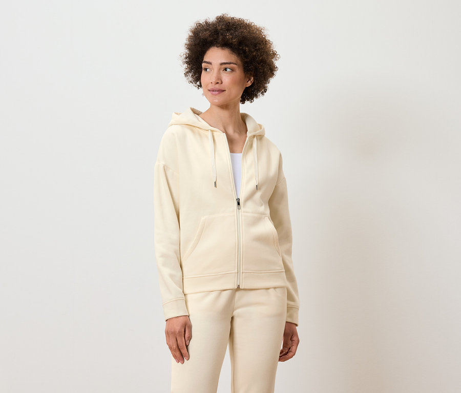 Kıvırcık saçlı kadın, krem rengi, fermuarlı kapüşonlu sweatshirt ve eşofman altıyla poz veriyor.