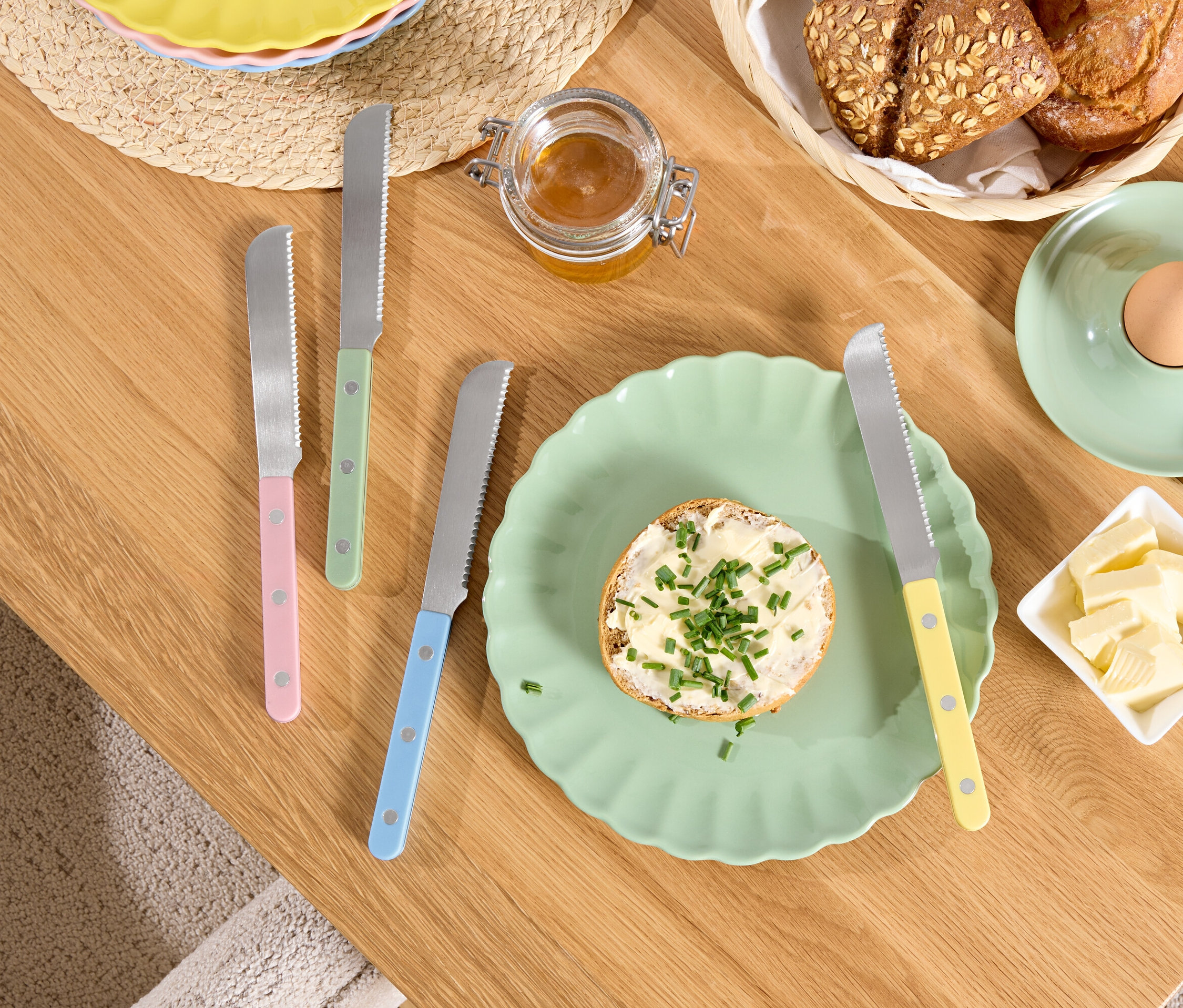 Ahşap bir masanın üzerinde tereyağlı ve otlu bir ekmek tabağı, bir kasede tereyağı, bir yumurtalıkta bir yumurta ve renkli saplı bıçaklar bulunmaktadır.