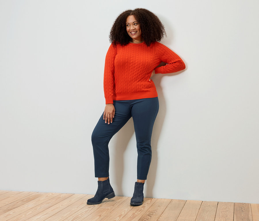 Kıvırcık saçlı gülümseyen bir kadın, turuncu bir kazak, mavi pantolon ve botlarla poz veriyor.