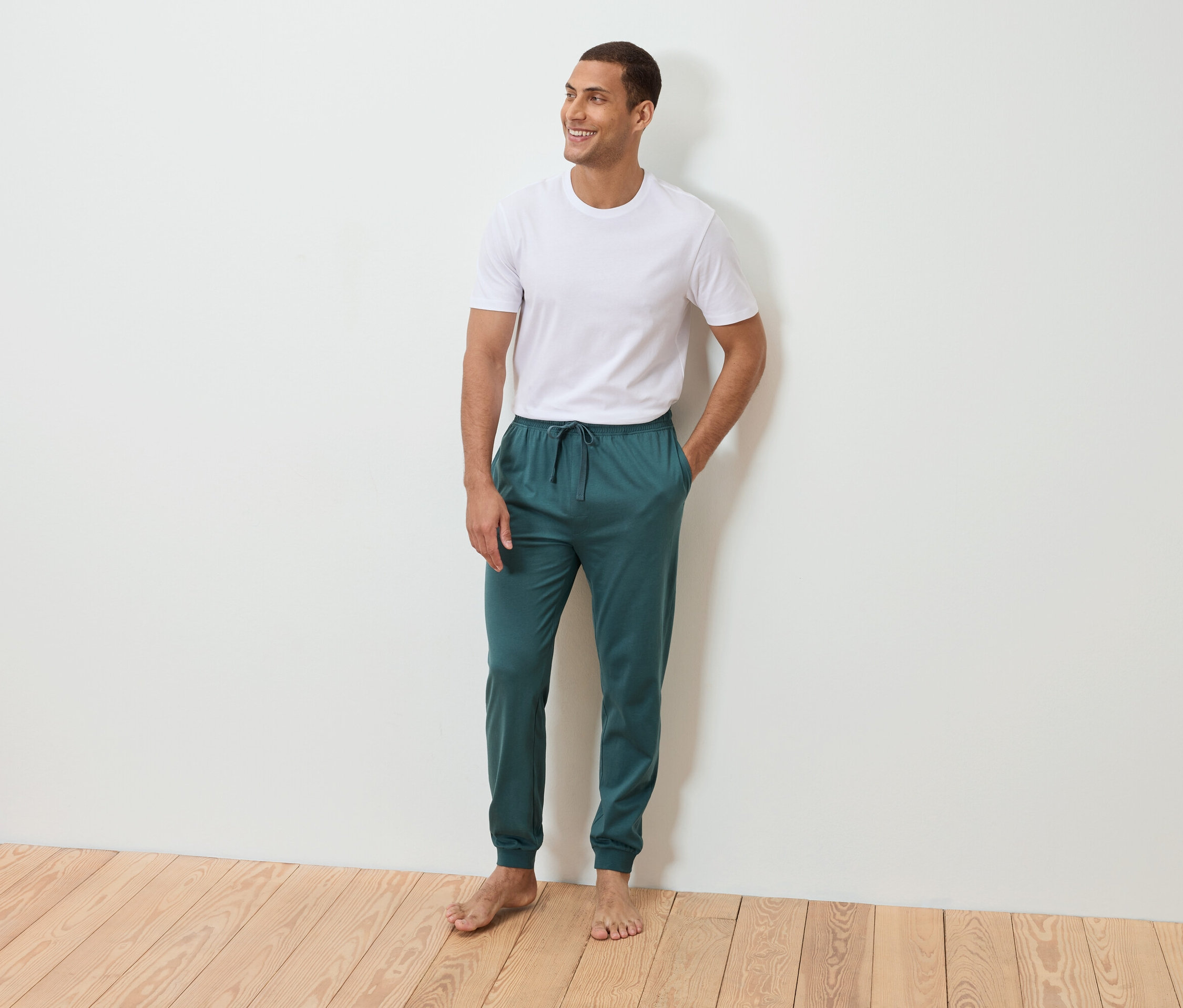 Bir adam beyaz bir duvarın önünde, eli cebinde, çıplak ayakla duruyor. Beyaz bir tişört ve petrol rengi eşofman altı giyiyor.