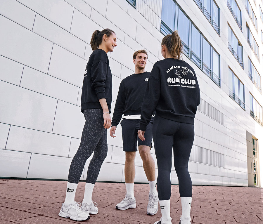 Üç kişi siyah sweatshirt ve spor kıyafetleriyle sohbet ediyor. Bir kadın gri-siyah Spor Tayt "ActivePower" ve Unisex Coffee Crew Tenis Çorapları giyiyor. Bir erkek siyah Spor Şortu ve beyaz çorap giyiyor. Diğer kadın siyah Spor Tayt "ActivePower" ve arkasında "RUN CLUB" logolu sweatshirt giyiyor.