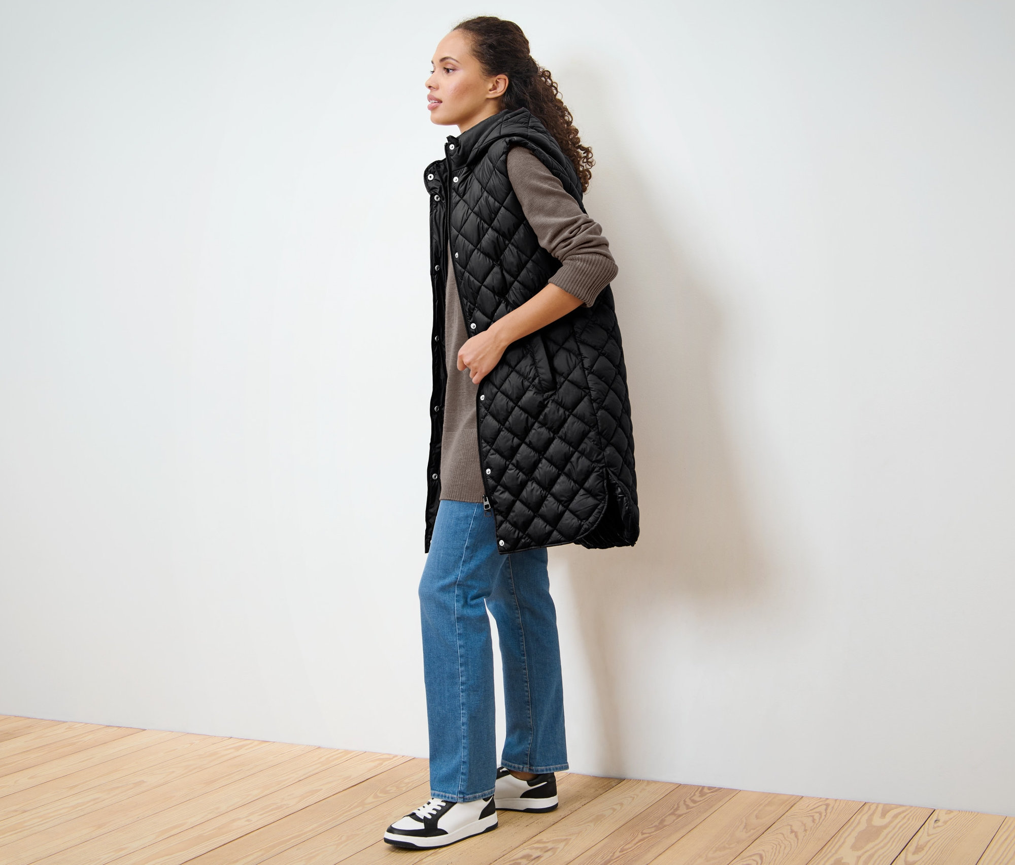 Kadın yan duruyor ve kapüşonlu uzun bir kapitone yelek giyiyor.