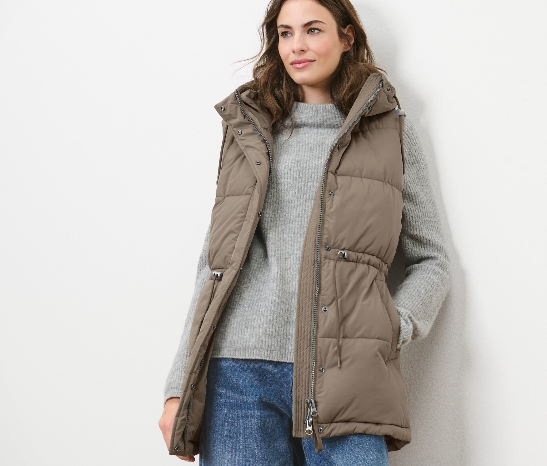 Kadın kapüşonlu kapitone yelek, kazak ve kot pantolon giyiyor.