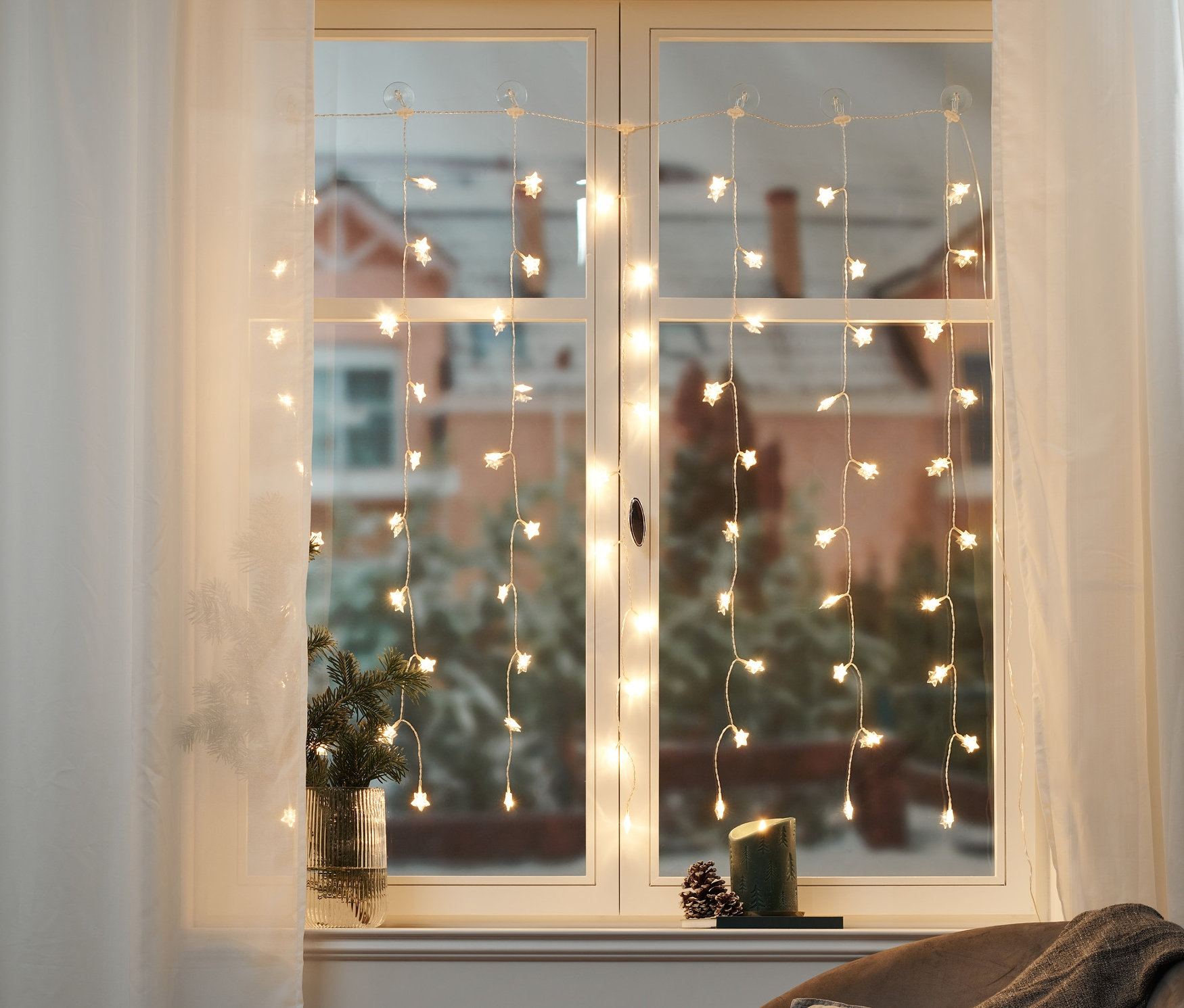 Pencerede LED Yıldız Işık Perdesi asılı duruyor. Odada bir koltuk, pencere pervazında çam dalları olan bir vazo ve bir mum var.