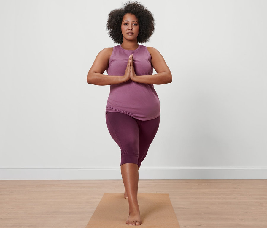 Kadın mürdüm rengi 3/4 yoga pantolonu ve eflatun spor üstüyle yoga matı üzerinde duruyor.