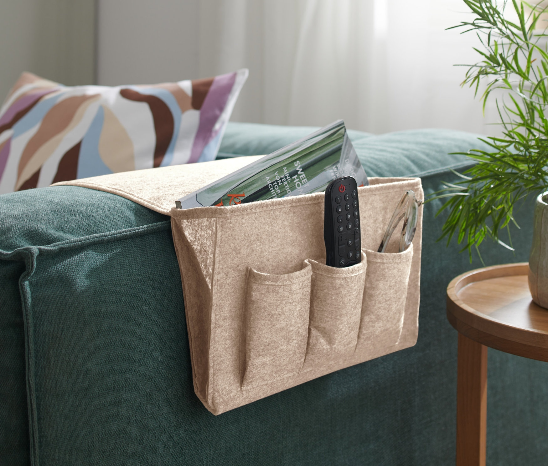 Bej rengi Yatak ve Koltuk Kenarı Düzenleyici, yeşil bir kanepenin kol dayanağında asılı duruyor, içinde bir uzaktan kumanda ve bir dergi var.