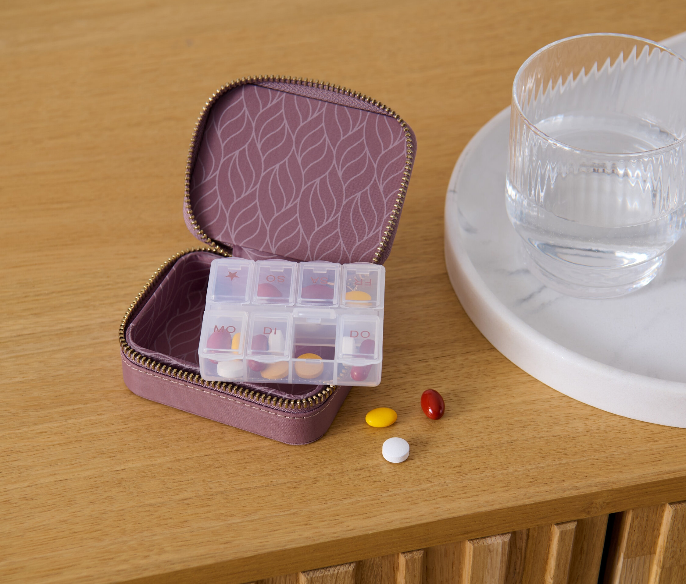 Açık mor renkli, desenli iç yüzeye sahip ilaç kutusu, yanında bir bardak su ve ahşap bir masa üzerinde üç adet hap bulunuyor.