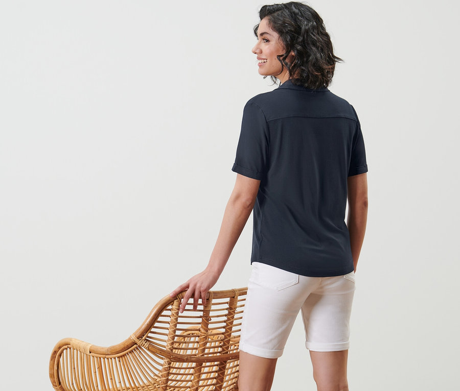 Kadın hasır bir sandalyenin yanında duruyor ve lacivert, düğmeli bir bluz giyiyor.