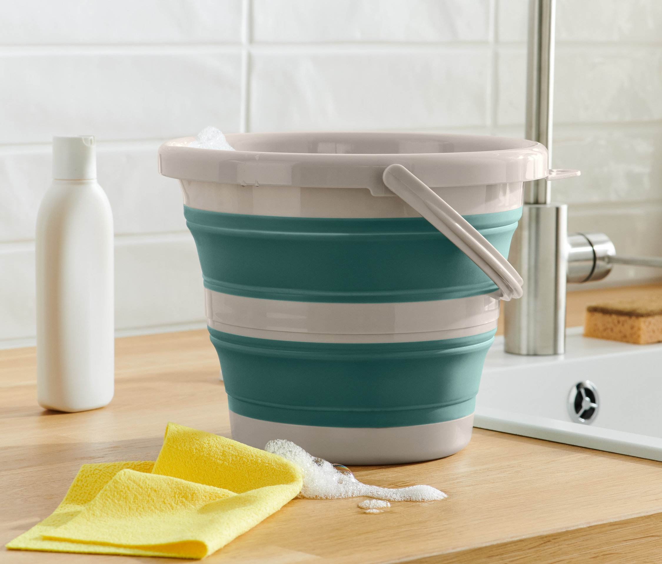 Katlanabilen temizlik kovası, sarı bir bezin ve bir şişenin yanında duruyor. Arka planda bir lavabo var.