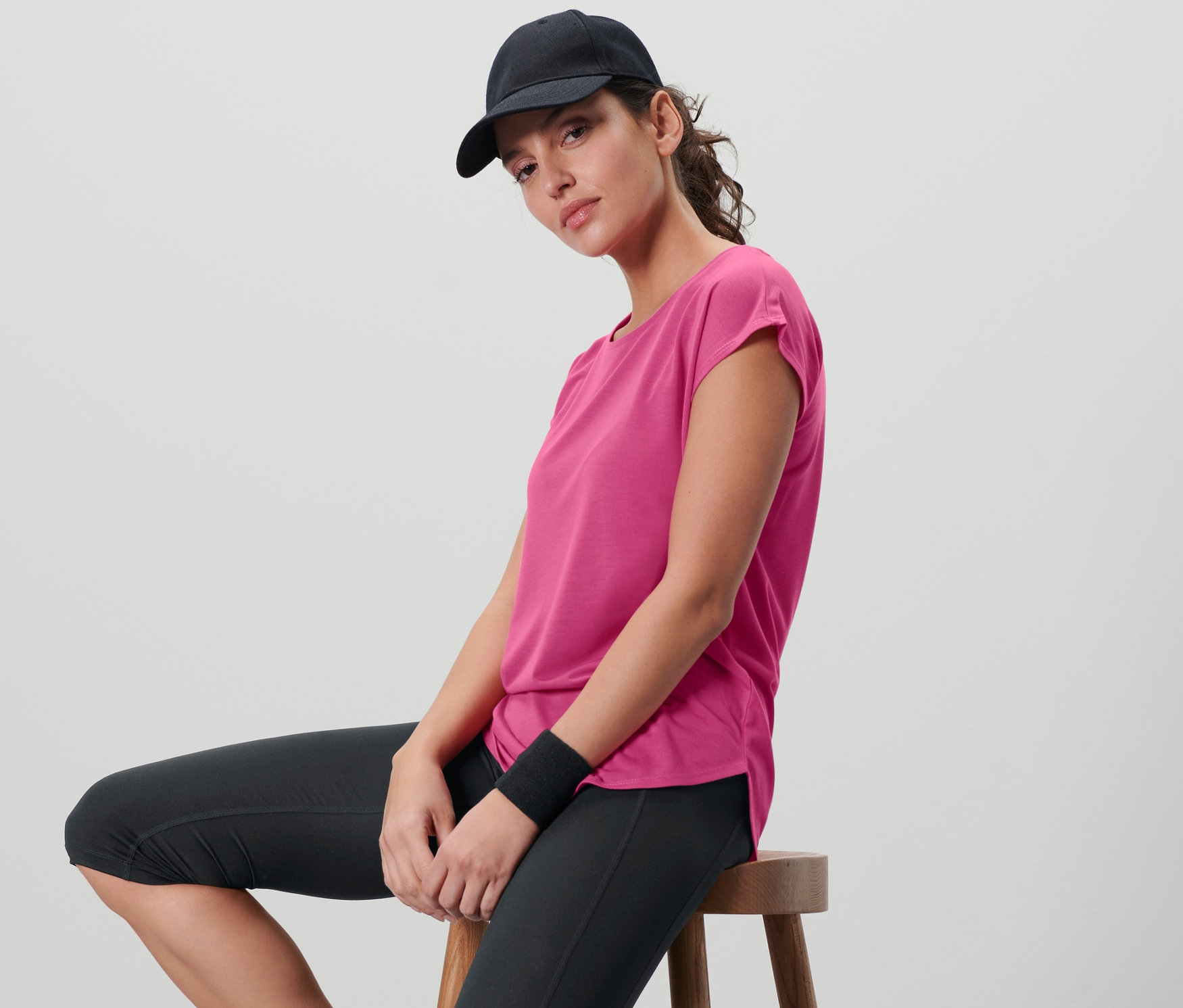 Kadın bir taburede oturuyor ve fuşya rengi bir spor tişörtü, siyah spor taytı ve siyah bir beyzbol şapkası giyiyor.