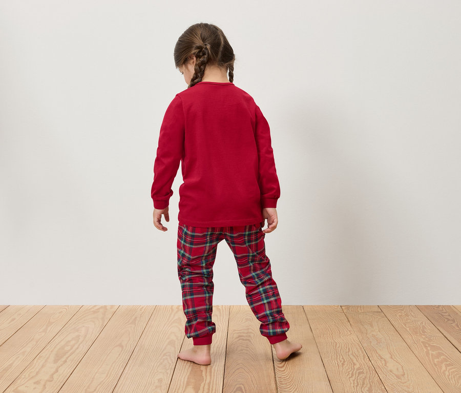 Örgülü saçlı kız, kırmızı, uzun kollu pijaması ve ekose pantolonuyla ahşap zeminde izleyiciye arkası dönük duruyor.