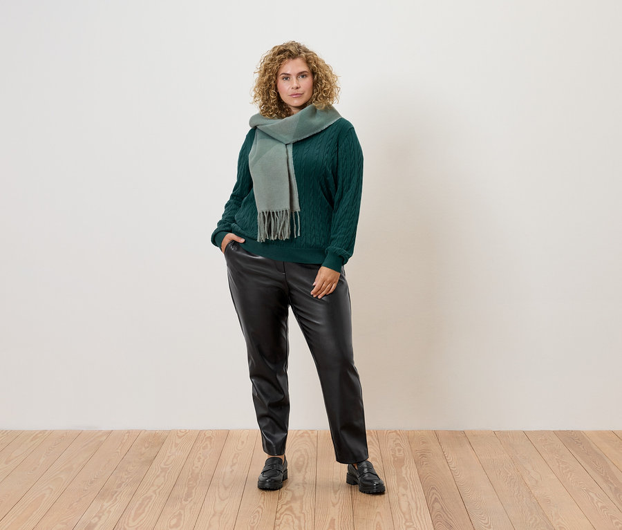 Kıvırcık saçlı bir kadın yeşil bir kazak, eşarp ve siyah deri pantolonla poz veriyor.
