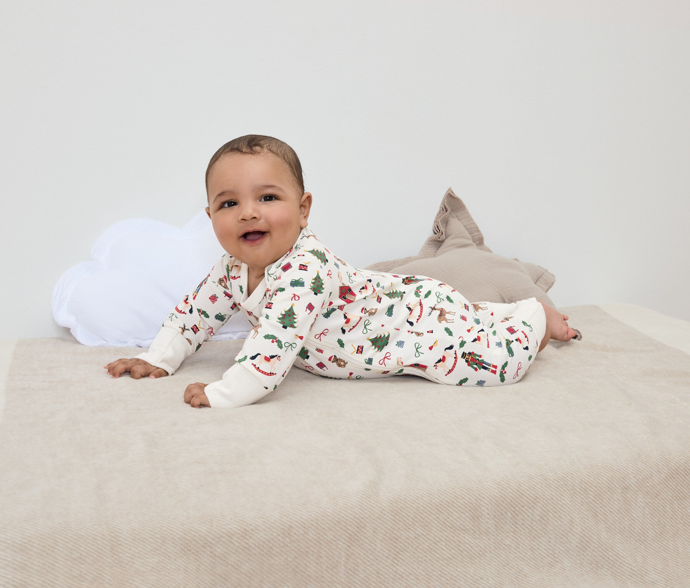 Sırıtan bir bebek, Noel desenli bir battaniyenin üzerinde, beyaz bulut şeklinde bir yastık ve kahverengi yıldız şeklinde bir yastığın yanında yüzüstü yatıyor.