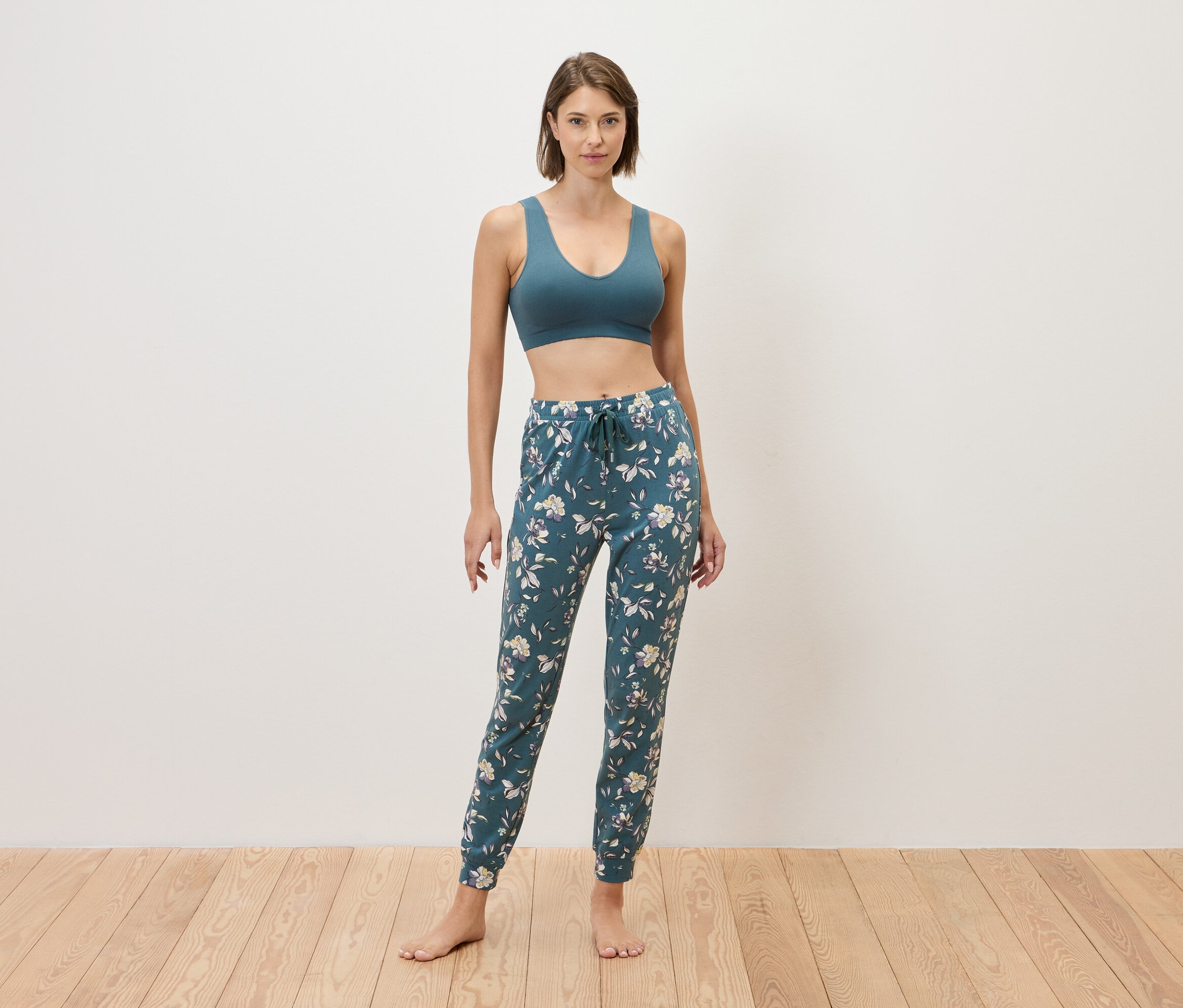 Kadın çıplak ayakla bir duvarın önünde duruyor, mavi bir spor sütyeni ve çiçek desenli mavi eşofman altı giyiyor.